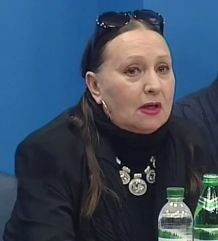 Лариса Кадочникова в чёрной одежде с украшением сидит у стола